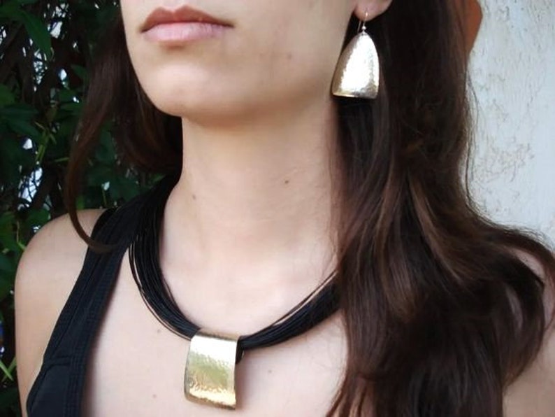 May include: A woman wearing a black choker necklace with a large, rectangular pendant. The pendant is made of hammered metal and has a gold finish. The woman is also wearing silver earrings with a hammered metal design.