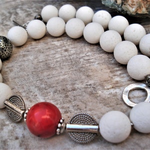 May include: A white lava rock beaded necklace with silver accents, including a round pendant, a small bell, and a large round bead with a textured surface. The necklace also features a red coral bead.
