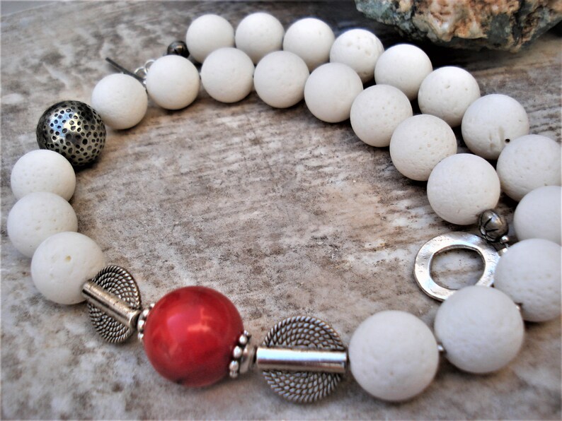 May include: A white lava rock beaded necklace with silver accents, including a round pendant, a small bell, and a large round bead with a textured surface. The necklace also features a red coral bead.
