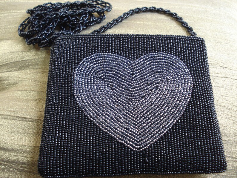Puede incluir: Un bolso de noche negro de cuentas con un dise&ntilde;o de coraz&oacute;n plateado. El bolso tiene una correa de cadena de cuentas.