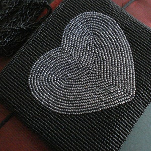 Puede incluir: Un bolso de mano negro de cuentas con un dise&ntilde;o de coraz&oacute;n plateado.