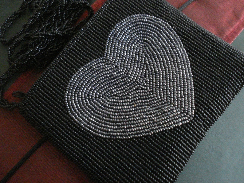 Puede incluir: Un bolso de mano negro de cuentas con un dise&ntilde;o de coraz&oacute;n plateado.