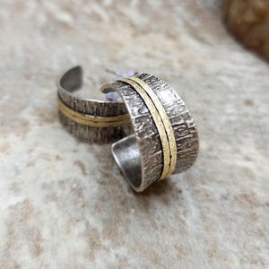 May include: A pair of silver hoop earrings with a textured finish and a gold band. The earrings have a rustic, handcrafted appearance. The jewellery is displayed on a light-coloured surface.