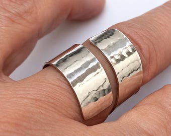 May include: Two wide, silver-colored rings with a hammered, textured finish. The bands have a reflective, metallic surface and are worn together on a finger, showcasing a curved, open-ended design with a polished, shiny appearance.