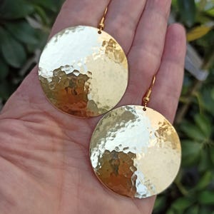 以下が含まれることがあります： 金色のハンマー加工された金属製のディスクイヤリングのペア。イヤリングは丸く、テクスチャ加工された表面とフック留めが付いています。イヤリングは、緑の葉の背景に対して手で持たれています。