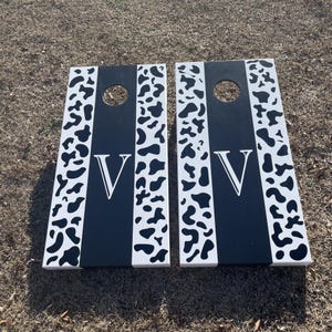 May include: Two black and white wooden cornhole boards with a cow print design and the letter "V" painted on each board.