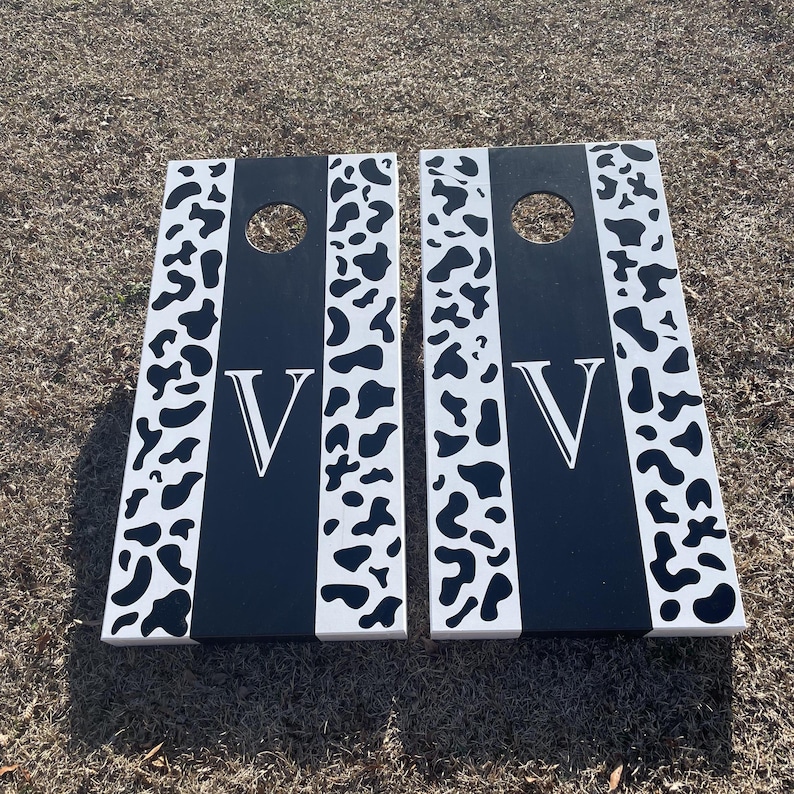 May include: Two black and white wooden cornhole boards with a cow print design and the letter "V" painted on each board.
