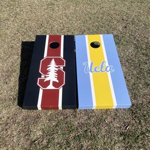 May include: Two wooden cornhole boards with team logos. The left board is black with a red stripe and a white Stanford tree logo. The right board is blue with a yellow stripe and the word "UCLA" in white.