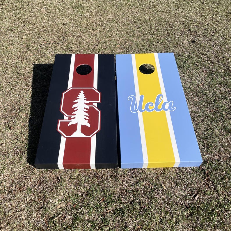 May include: Two wooden cornhole boards with team logos. The left board is black with a red stripe and a white Stanford tree logo. The right board is blue with a yellow stripe and the word "UCLA" in white.