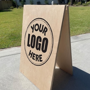 May include: A-frame wooden sign with the text "YOUR LOGO HERE" in a black circular design. The sign is made of light-colored wood and stands on a concrete surface, ideal for businesses to display their branding.