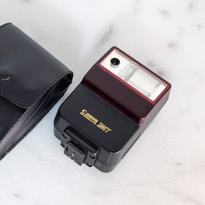 May include: A black Canon Speedlite 244T flash unit with a matching black leather case. The flash has a red top and a silver flash head. The case has the Canon logo embossed on it.