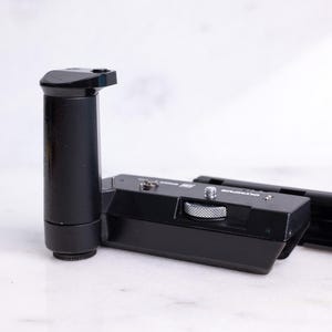 Puede incluir: Un agarre de cámara negro con un mango cilíndrico y una base rectangular. El agarre tiene un dial plateado y una extensión rectangular negra. Los artículos están sobre una superficie blanca.