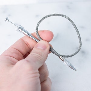May include: A silver flexible shaft tool with a small, pointed tip. The shaft is encased in a braided, silver-colored cable.