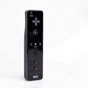 Puede incluir: Un mando a distancia Wii negro con un acabado brillante. El mando tiene un botón de encendido, un pad direccional, un botón A, un botón de inicio y botones numéricos. La palabra "Wii" está impresa en la parte inferior.