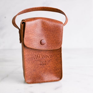 May include: A brown leather camera case with a strap and a button closure. The case has the text "Nikon S57 Japan" embossed on the front.