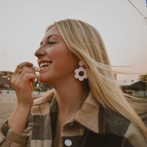 May include: A person applying lip balm. The person is wearing a plaid jacket and flower-shaped earrings. The earrings are light pink with a dark pink center. A gold ring is on the person's finger.