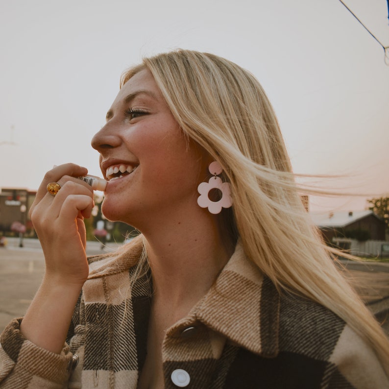 May include: A person applying lip balm. The person is wearing a plaid jacket and flower-shaped earrings. The earrings are light pink with a dark pink center. A gold ring is on the person's finger.