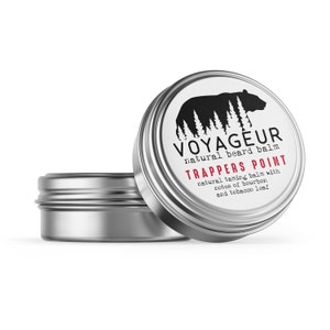 Op de afbeelding: Twee zilveren blikken baardbalsem. Eén blik is open en onthult het product. Het etiket toont een zwart berensilhouet boven de tekst "VOYAGEUR natural beard balm" en "TRAPPERS POINT" in rood. De tekst hieronder luidt "natural taming balm with notes of bourbon and tobacco leaf."