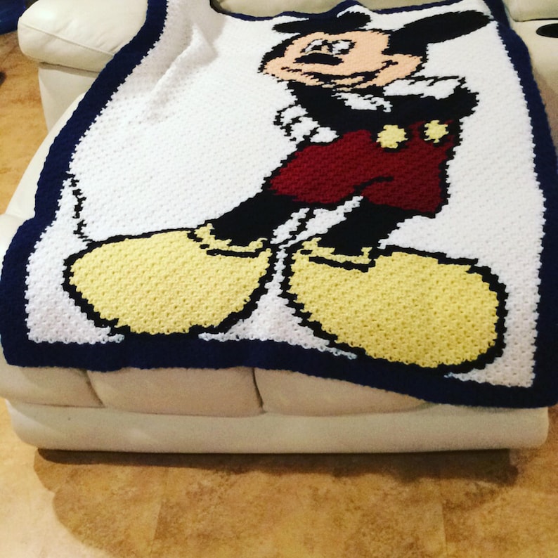 May include: A white crocheted blanket with a blue border featuring a cartoon image of Mickey Mouse. Mickey is wearing red shorts and yellow shoes.