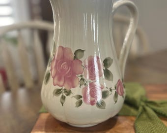 May include: A white ceramic jug with a floral design featuring pink roses and green leaves. The jug has a handle and a wide mouth.