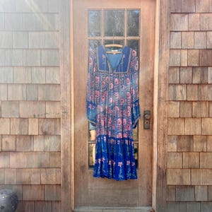 Puede incluir: Un vestido largo azul con un estampado floral de flores rosas y hojas verdes cuelga de una percha de madera. El vestido tiene un escote cuadrado y una falda escalonada. El vestido está colgado de una puerta de madera.