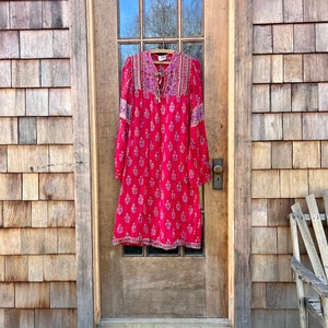 May include: A red, long-sleeved dress with a floral print and purple trim. The dress is hanging on a hanger in front of a wooden door.