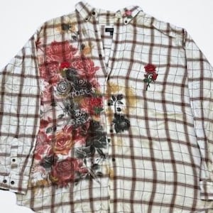 Può includere: Una camicia di flanella a quadri bianchi e marroni con una stampa floreale sul lato sinistro. La stampa floreale presenta rose rosse, bianche e nere. La camicia ha un colletto abbottonato e maniche lunghe.