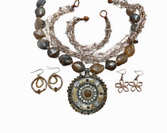 Könnte beinhalten: Ein braunes und silbernes Halsketten-Set mit einem großen runden Anhänger, braunen Steinperlen und einem passenden Paar Ohrringen. Der Anhänger hat ein kreisförmiges Design mit einem kleineren braunen Stein in der Mitte.