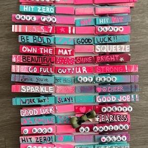 Può includere: Una collezione di mollette decorative in legno, dipinte in tonalità rosa e verde acqua, con varie frasi motivazionali e parole come "SLAY" e "BEAUTIFUL" scritte sopra. Alcune mollette hanno dettagli glitterati e piccoli abbellimenti.