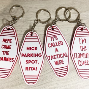 Op de afbeelding: Vier witte motel sleutelhangers met rode strepen en rode tekst. De sleutelhangers zeggen "Here Come The Grannies", "Nice Parking Spot, Rita!", "It's Called A Tactical Wee", en "I'm The Flamingo Queen".
