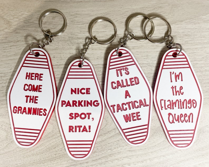 Op de afbeelding: Vier witte motel sleutelhangers met rode strepen en rode tekst. De sleutelhangers zeggen "Here Come The Grannies", "Nice Parking Spot, Rita!", "It's Called A Tactical Wee", en "I'm The Flamingo Queen".
