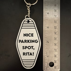 Op de afbeelding: Witte motel sleutelhanger met zwarte tekst "NICE PARKING SPOT, RITA!" hangend aan een zilveren sleutelring.