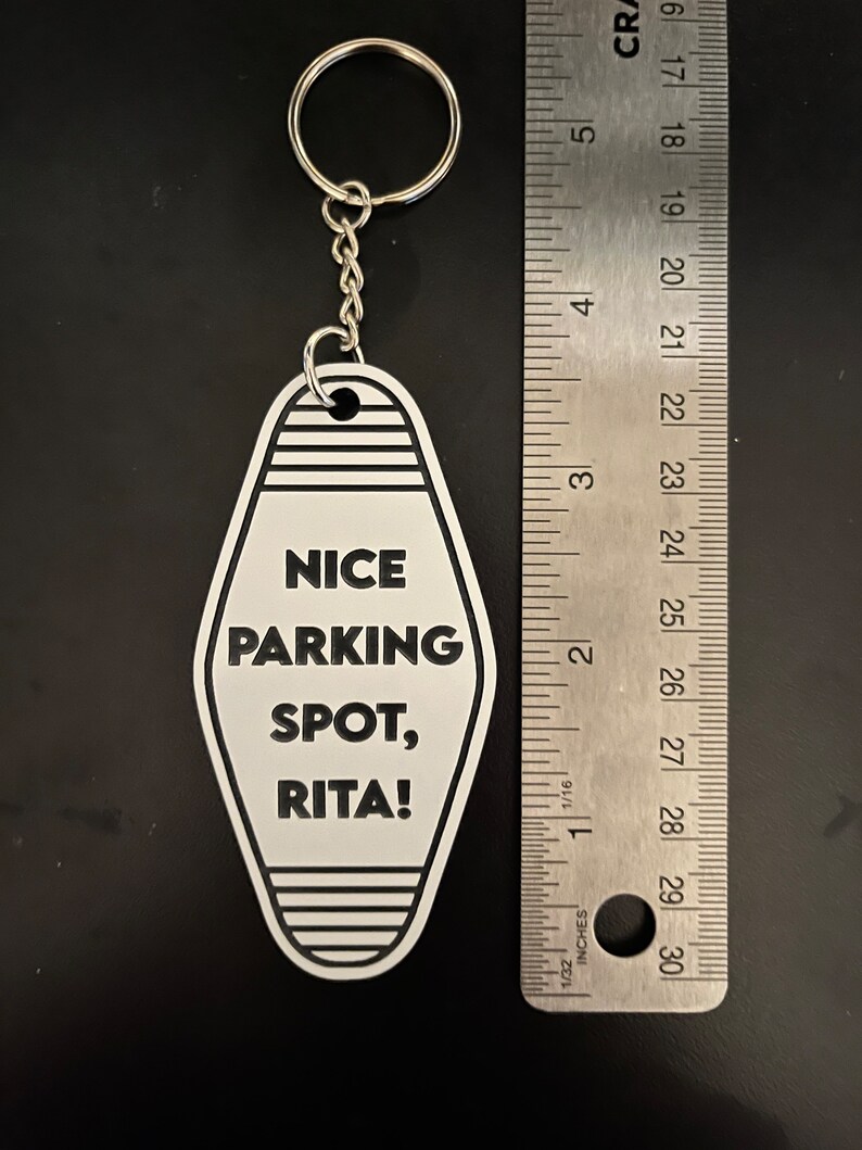 Op de afbeelding: Witte motel sleutelhanger met zwarte tekst "NICE PARKING SPOT, RITA!" hangend aan een zilveren sleutelring.