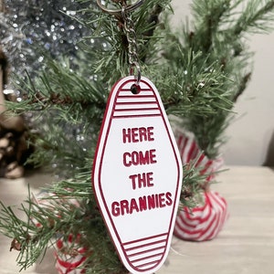 Op de afbeelding: Witte motel sleutelhanger met rode rand en de tekst "Here Come The Grannies" hangend aan een zilveren ketting aan een kleine kunstkerstboom.
