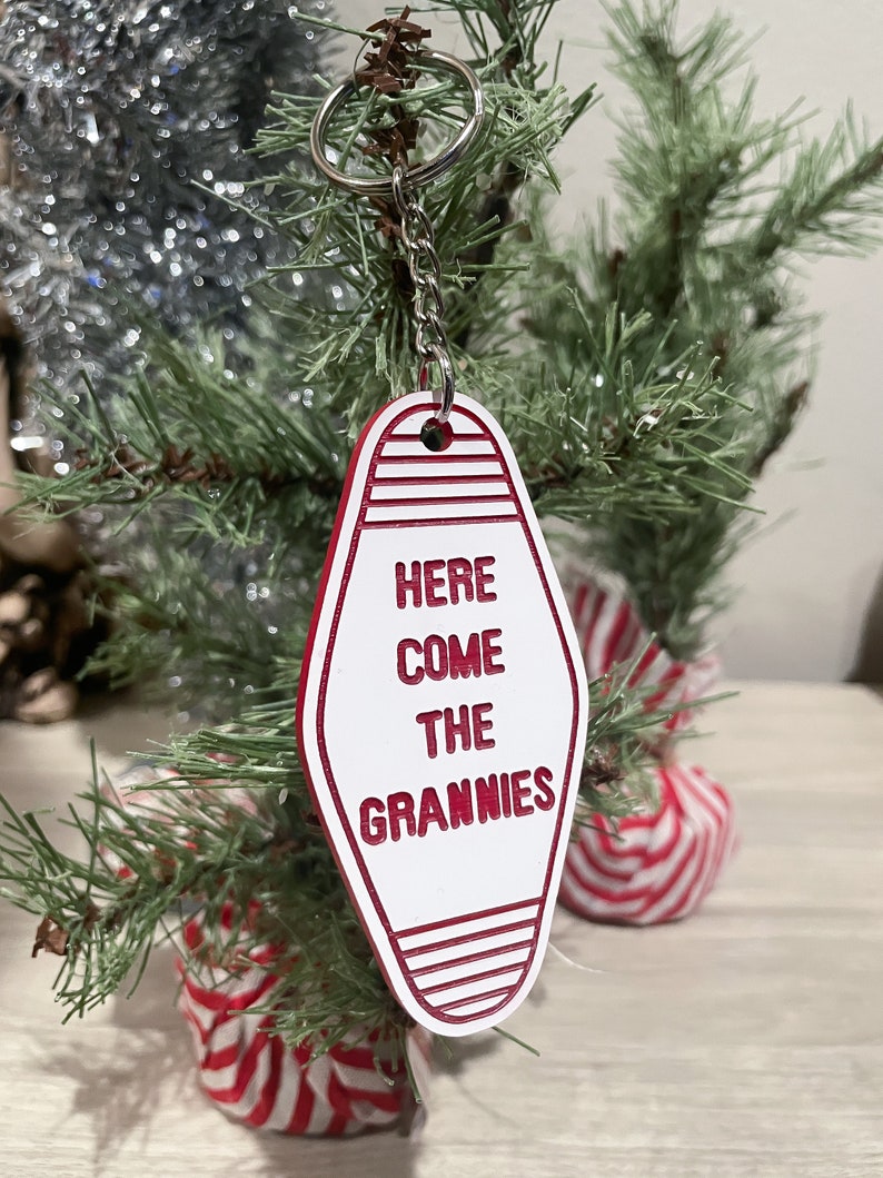 Op de afbeelding: Witte motel sleutelhanger met rode rand en de tekst "Here Come The Grannies" hangend aan een zilveren ketting aan een kleine kunstkerstboom.