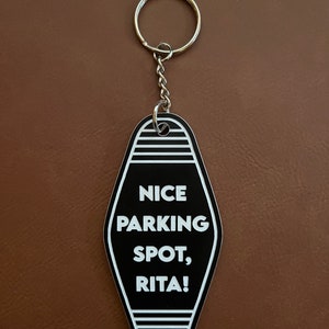 Op de afbeelding: Zwart-witte motel sleutelhanger met de tekst "NICE PARKING SPOT, RITA!"