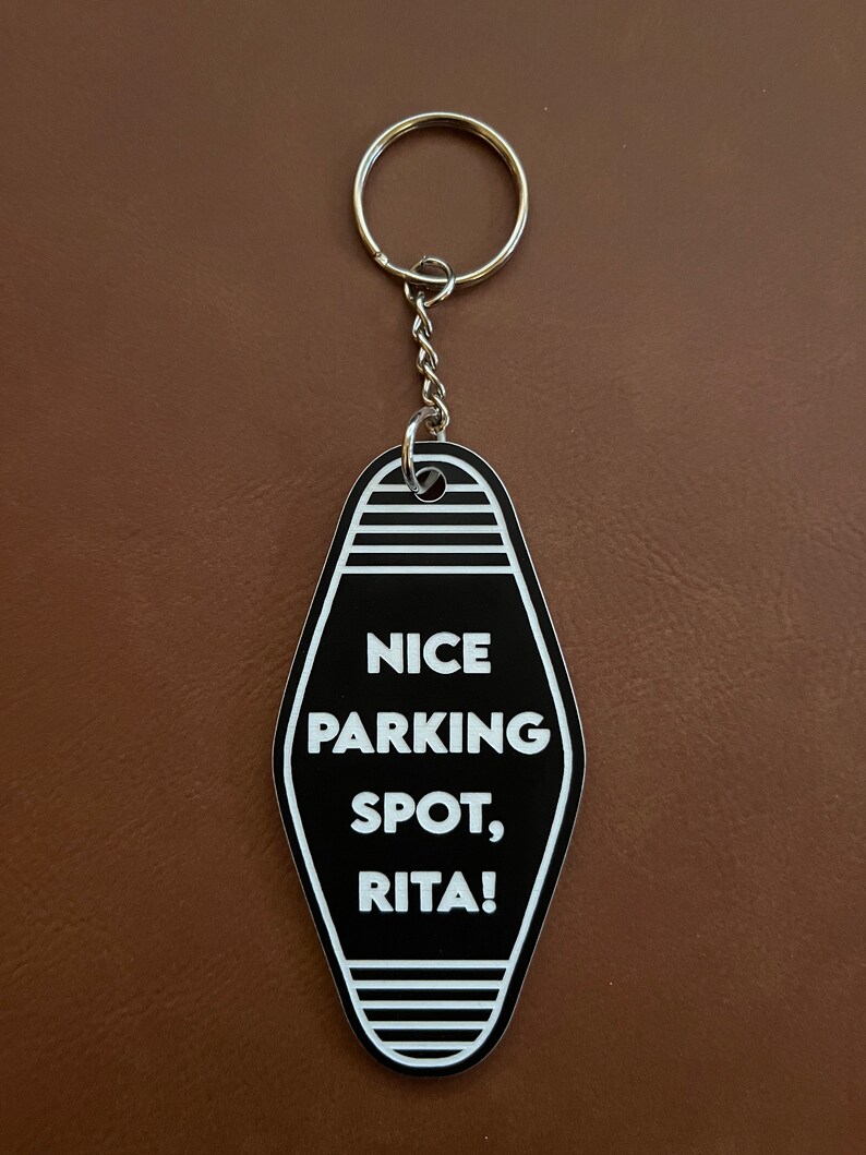 Op de afbeelding: Zwart-witte motel sleutelhanger met de tekst "NICE PARKING SPOT, RITA!"