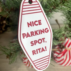 Op de afbeelding: Witte motel sleutelhanger ornament met rode strepen en de tekst "NICE PARKING SPOT, RITA!" hangend aan een kerstboom.