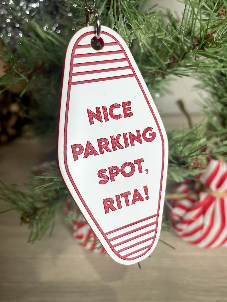 Op de afbeelding: Witte motel sleutelhanger ornament met rode strepen en de tekst "NICE PARKING SPOT, RITA!" hangend aan een kerstboom.