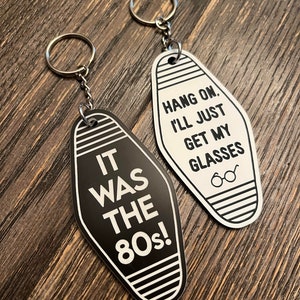 Op de afbeelding: Twee zwart-witte motel sleutelhangers met een zilveren sleutelring. De ene sleutelhanger zegt "It was the 80s!" en de andere zegt "Hang on. I'll just get my glasses."