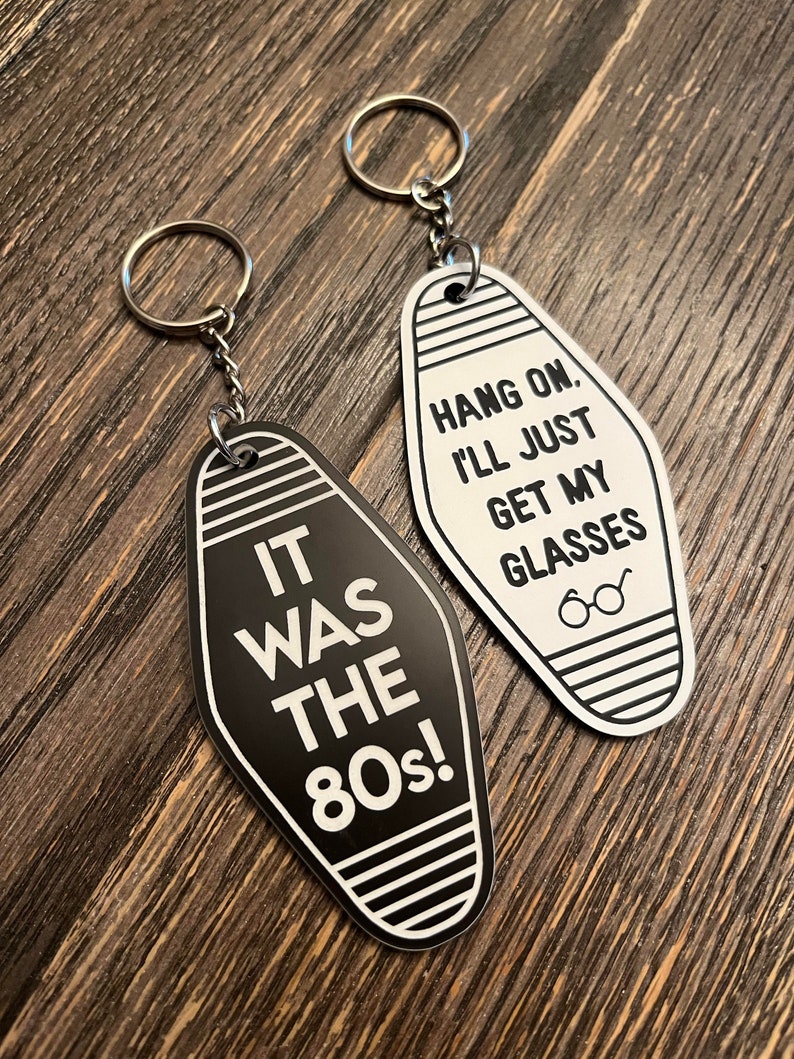 Op de afbeelding: Twee zwart-witte motel sleutelhangers met een zilveren sleutelring. De ene sleutelhanger zegt "It was the 80s!" en de andere zegt "Hang on. I'll just get my glasses."