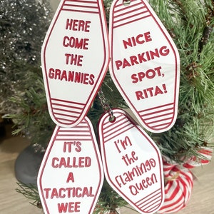 Op de afbeelding: Vier witte motel-sleutelhangers in de vorm van sleutels met rode rand en rode tekst. De hangers tonen de opschriften "Here Come The Grannies", "It's Called A Tactical Wee", "Nice Parking Spot, Rita!" en "I'm The Flamingo Queen".