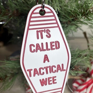 Op de afbeelding: Witte motel sleutelhanger ornament met rode tekst "It's called a tactical wee".