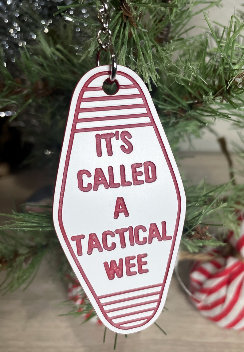 Op de afbeelding: Witte motel sleutelhanger ornament met rode tekst "It's called a tactical wee".