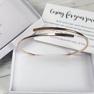 May include: A gold-toned bangle bracelet in a white box with a white card that reads "Caring for your jewellery. Keep jewellery away from moisture. Apply perfumes and lotions before wearing your jewellery. When sleeping, jewellery should be neatly stored. Not in use, jewellery should be neatly stored. Hepzibahpink.etsy.com"
