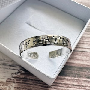 May include: A silver ring with an engraved design of a camper, trees, and mountains. The ring is in a white box.