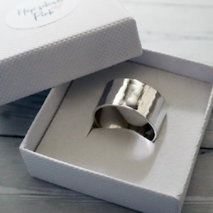 May include: A broad, polished silver ring is presented in a white textured gift box. The ring's surface is smooth and reflective. The box is open, displaying the ring's interior and the brand name "Hope & Pink" on a small sticker.