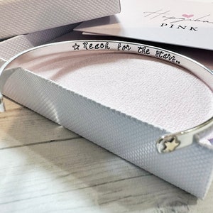 May include: A silver bangle bracelet with a star charm and the inscription "Reach for the stars..." The bracelet is in a white box with a pink heart logo.