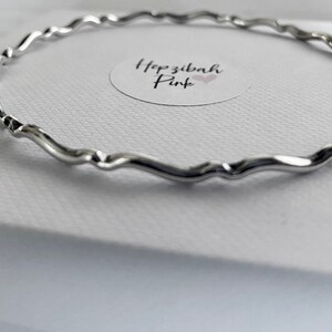 May include: A silver bangle bracelet with a wavy design. The bracelet is on a white surface with a small sticker that says "Hopzibah Pink".
