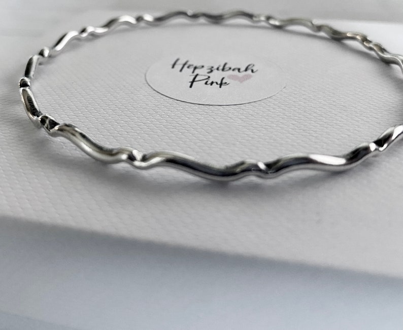 May include: A silver bangle bracelet with a wavy design. The bracelet is on a white surface with a small sticker that says "Hopzibah Pink".